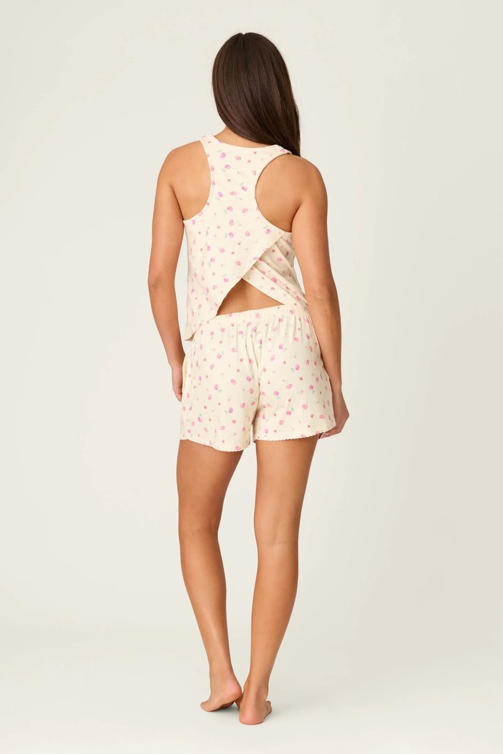 A woman stands barefoot, facing away, wearing the Zertz FOR THE MAMAS BLOOM PJ SHORT—a butter-yellow set with pink floral patterns. The racerback top features a crossover back and the loose shorts offer comfort for women&