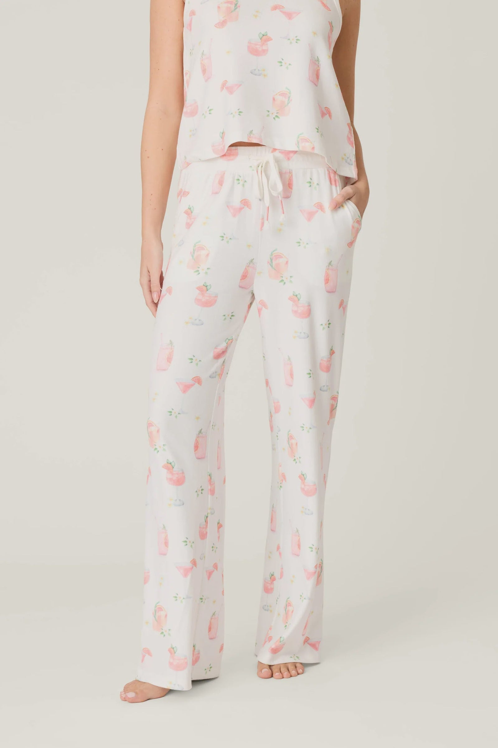 A person stands barefoot in Zertz's PALOMA MAMA PJ PANT and a matching sleeveless top, both made from buttery jersey with a lively print of women in pink bathtubs and floral accents on an ivory background.