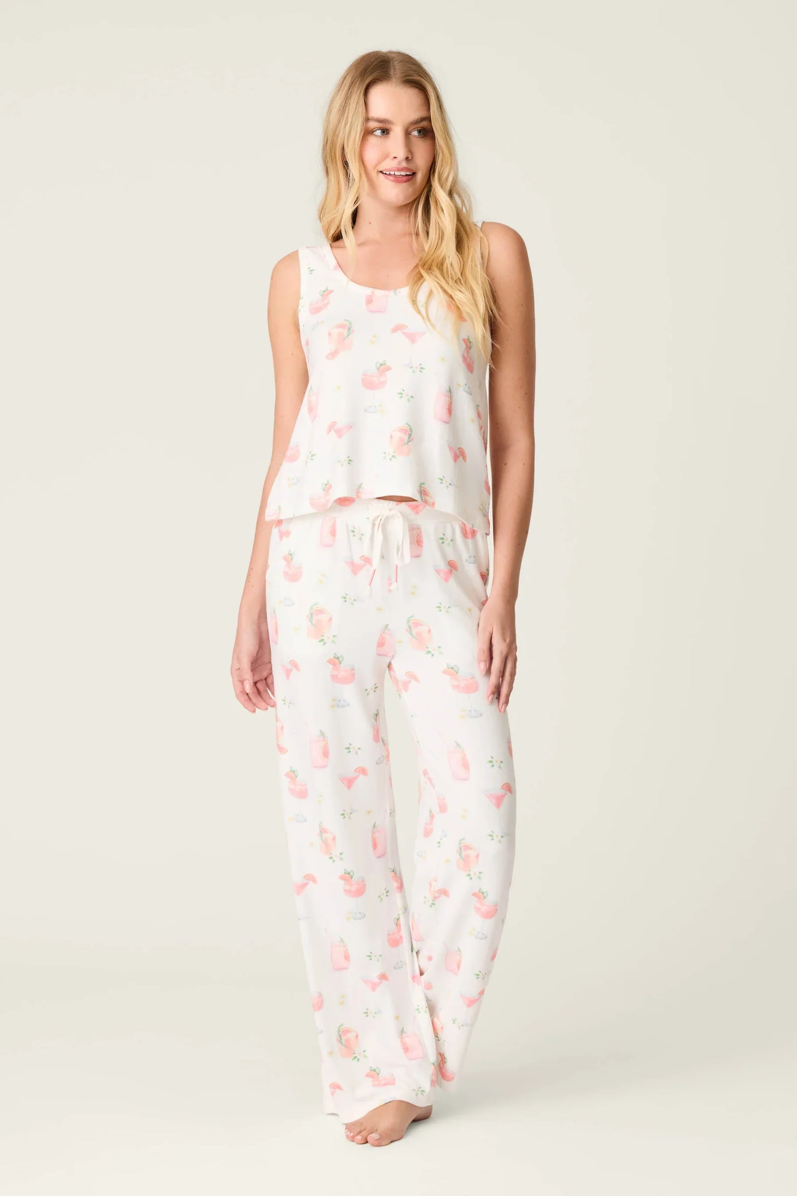 A woman with long blonde hair wears Zertz's PALOMA MAMA PJ PANT, a butter jersey pajama set with a sleeveless top and ivory pants in a light pink and peach floral print, standing barefoot against a plain backdrop.