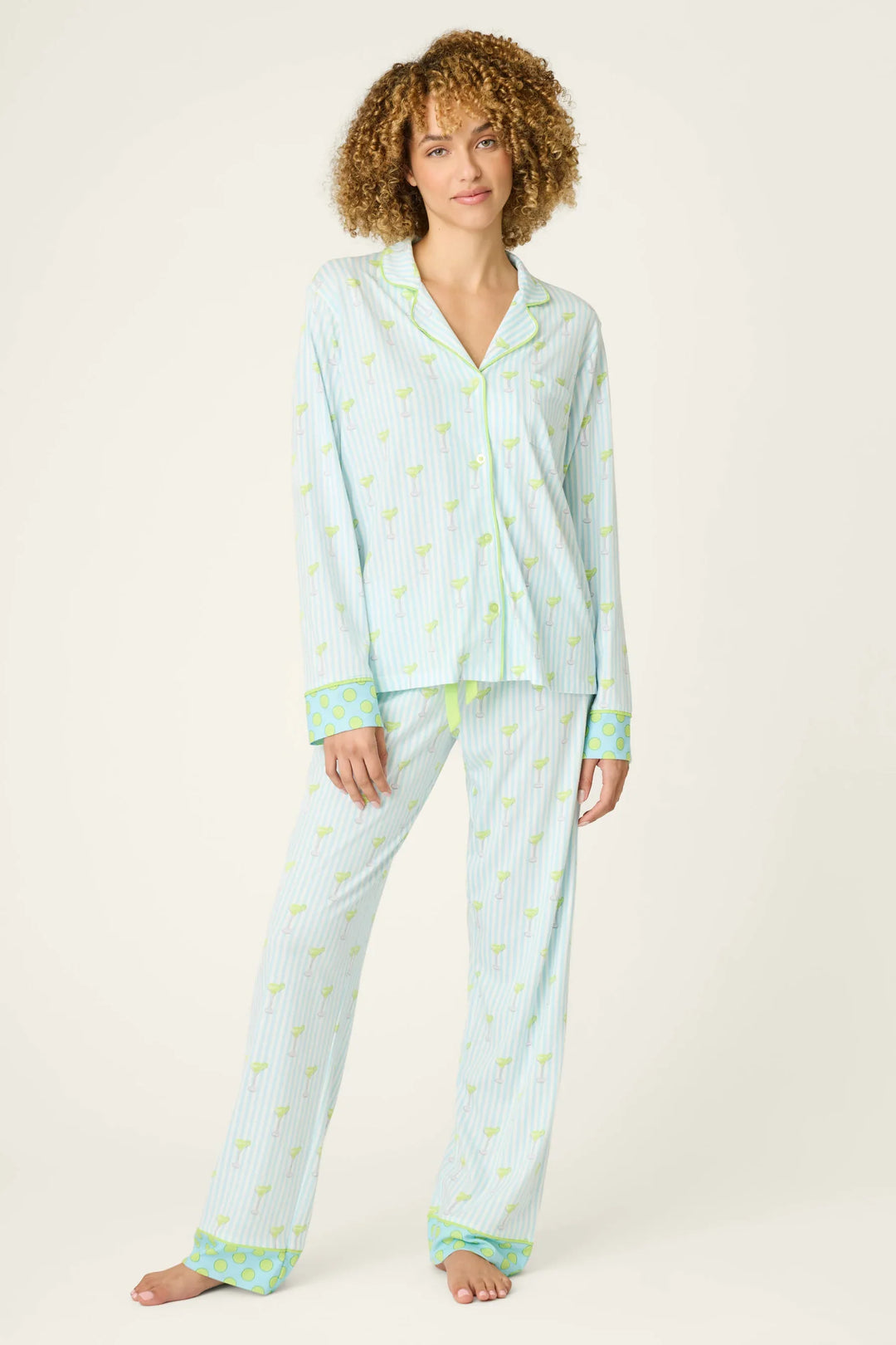 A person with curly hair wears the SALTY CADILLAC STRIPE LONG PJ SET by Zertz—light blue cocktail-inspired stripes with yellow and green pineapple prints and green cuffs—standing barefoot against a plain light background.