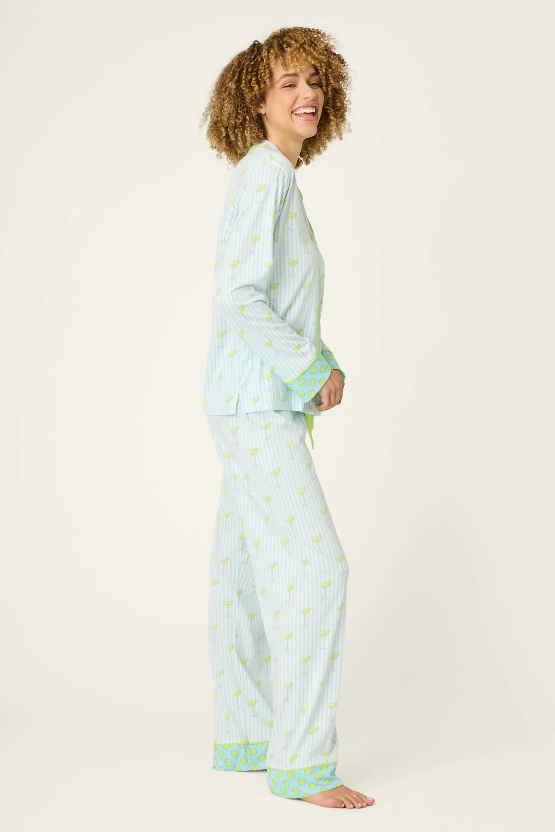 A woman with curly hair smiles while wearing the Zertz SALTY CADILLAC STRIPE LONG PJ SET, featuring blue-and-white stripes with green trim, standing barefoot against a plain background.