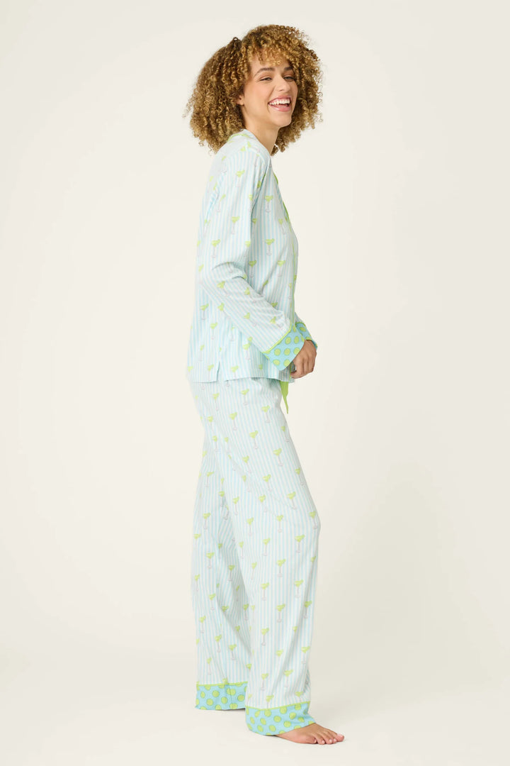A woman with curly hair smiles while wearing the Zertz SALTY CADILLAC STRIPE LONG PJ SET, featuring blue-and-white stripes with green trim, standing barefoot against a plain background.