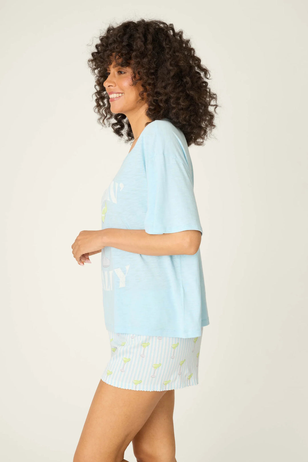 A woman with curly hair, smiling in profile, wears the Zertz S/S TEE PLAYFUL PRINT and striped shorts with a light pattern against a plain, light background.