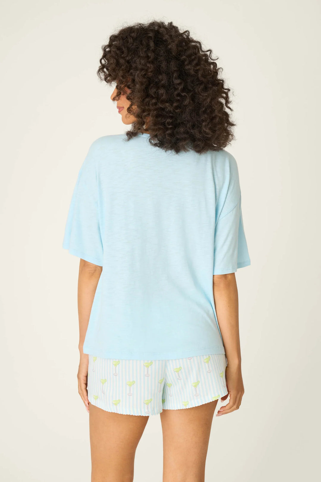 A person with curly hair stands facing away, wearing the Zertz S/S TEE PLAYFUL PRINT in light blue and white shorts featuring blue stripes and yellow lemon patterns, set against a plain light background.