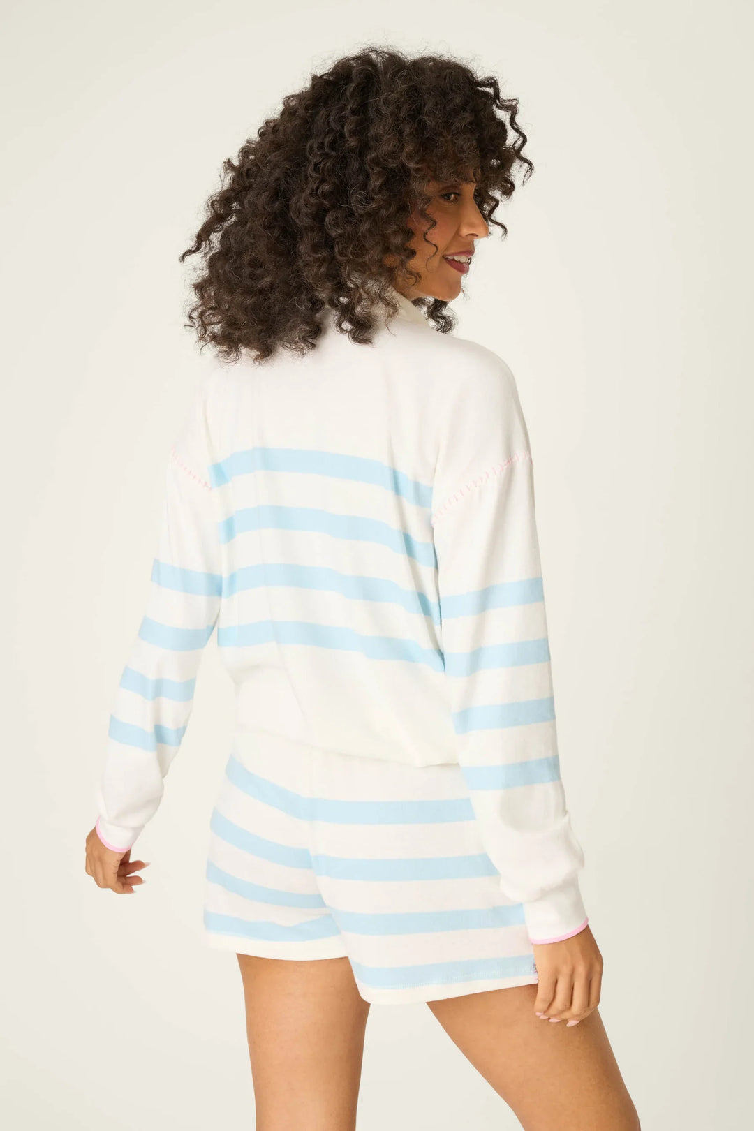 A person with curly hair smiles while wearing the Zertz Pupcation Palms Half Zip Pullover Sweater, featuring light blue stripes, and stands against a plain light background looking over their shoulder.
