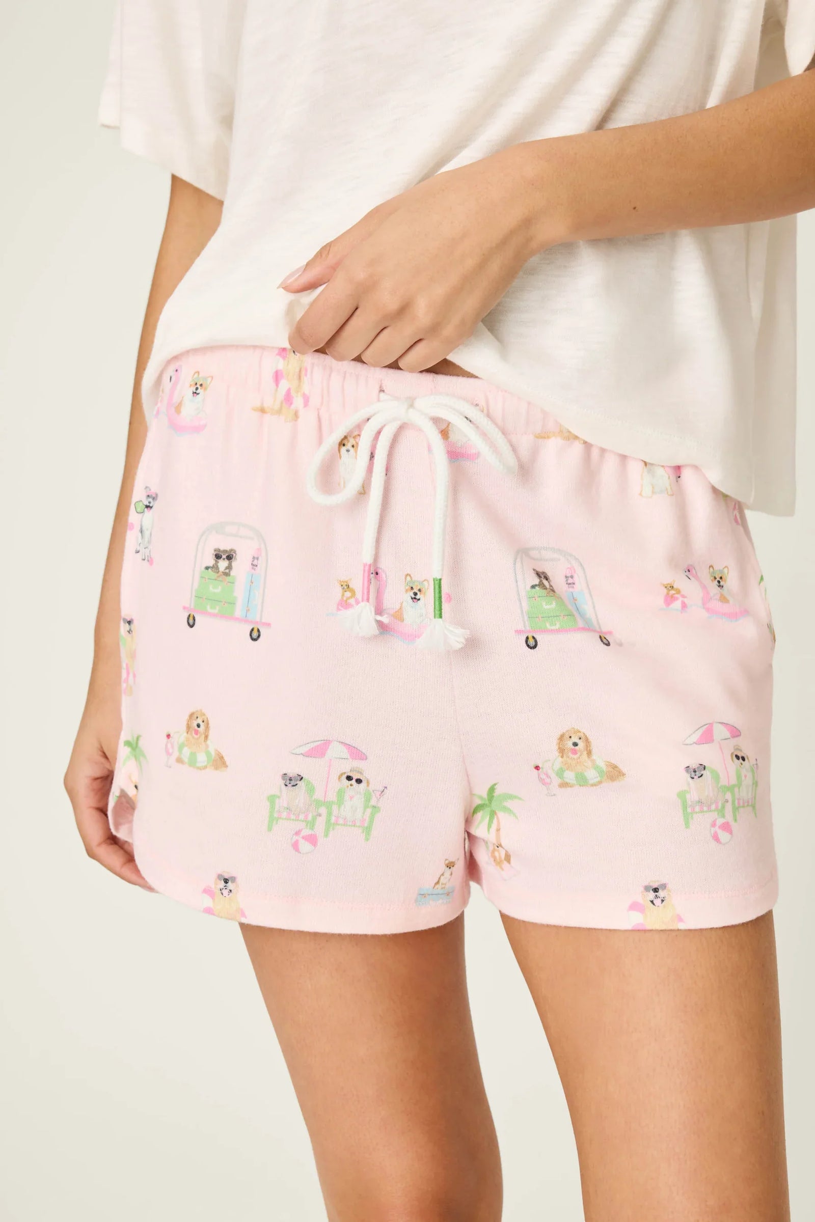 A person sports Zertz’s PUPCATION PALMS SUNNIES & SUNSHINE PJ SHORTS—runner-style with playful dog, surfboard, and beach prints—paired with a loose white tee for the perfect laid-back shore day look.