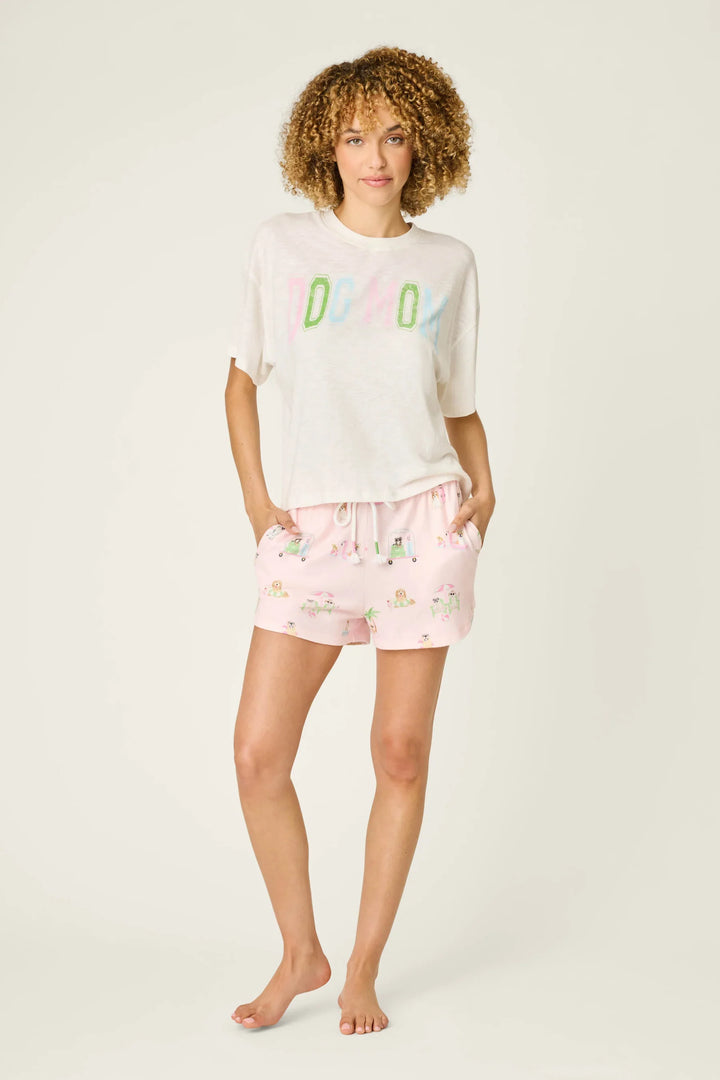 A woman with curly hair stands barefoot against a plain background, wearing a white DOG MOM t-shirt and Zertz PUPCATION PALMS SUNNIES &amp; SUNSHINE PJ SHORTS with a runner-style silhouette, her hands casually in her pockets.