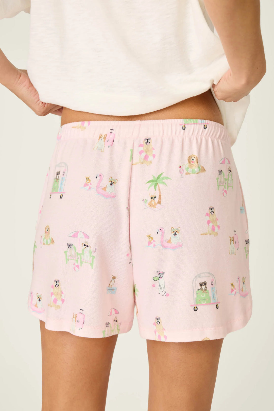 A person wears Zertz PUPCATION PALMS SUNNIES &amp; SUNSHINE PJ SHORTS—light pink lounge shorts with a playful dog-themed print, elastic waistband, and runner-style silhouette—paired with a white top for a relaxed vibe.