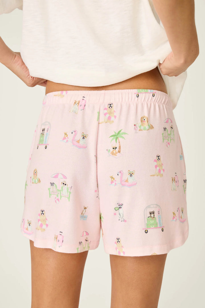 A person wears Zertz PUPCATION PALMS SUNNIES &amp; SUNSHINE PJ SHORTS—light pink lounge shorts with a playful dog-themed print, elastic waistband, and runner-style silhouette—paired with a white top for a relaxed vibe.