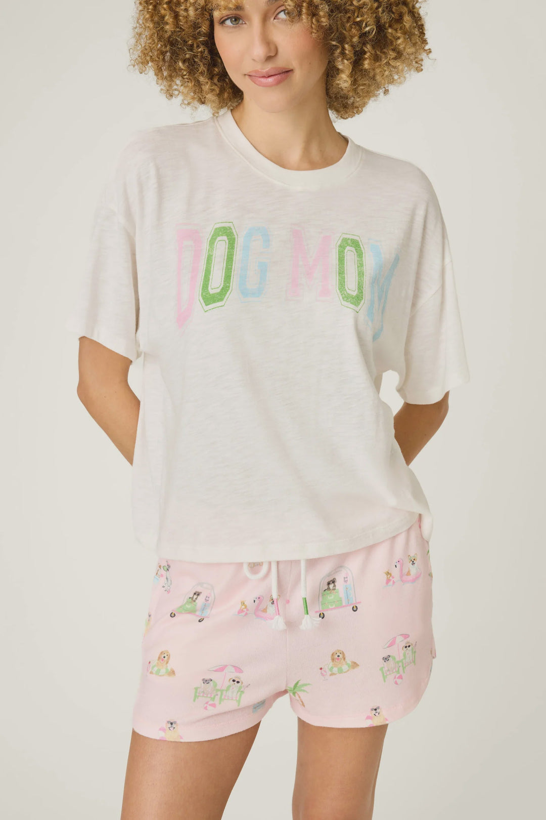 A woman with curly hair models the Zertz PUPCATION PALMS DOG MOM TEE in white, made from soft cotton-modal slub jersey, paired with pink dog-print pajama shorts, standing with her hands behind her back.