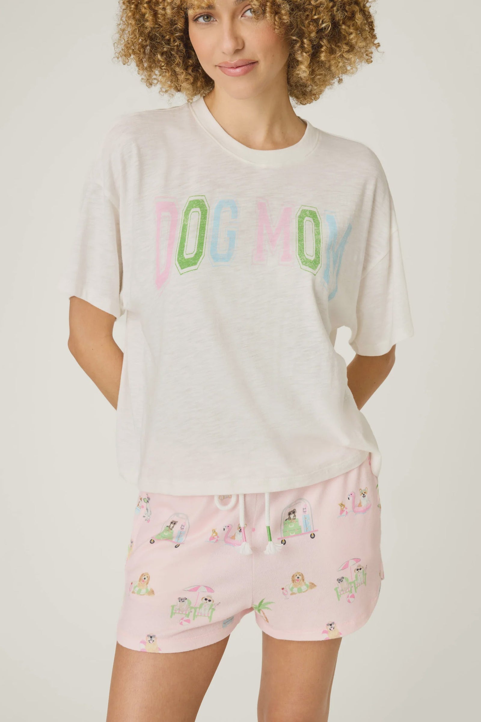 A woman with curly hair models the Zertz PUPCATION PALMS DOG MOM TEE in white, made from soft cotton-modal slub jersey, paired with pink dog-print pajama shorts, standing with her hands behind her back.