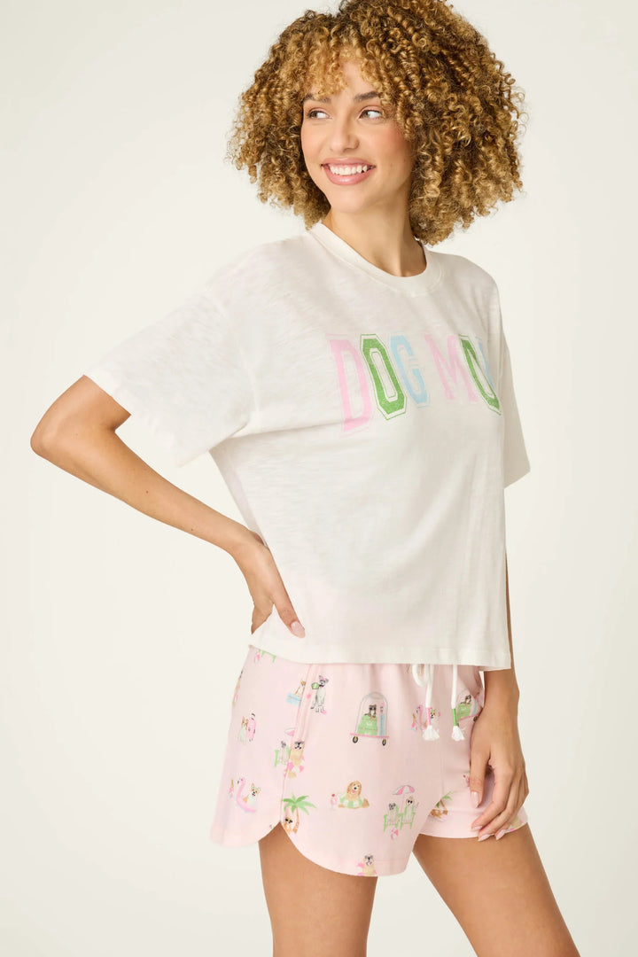 A woman with curly hair smiles, wearing the Zertz PUPCATION PALMS DOG MOM TEE featuring pastel lettering, paired with light pink shorts adorned with playful dog and camper prints.