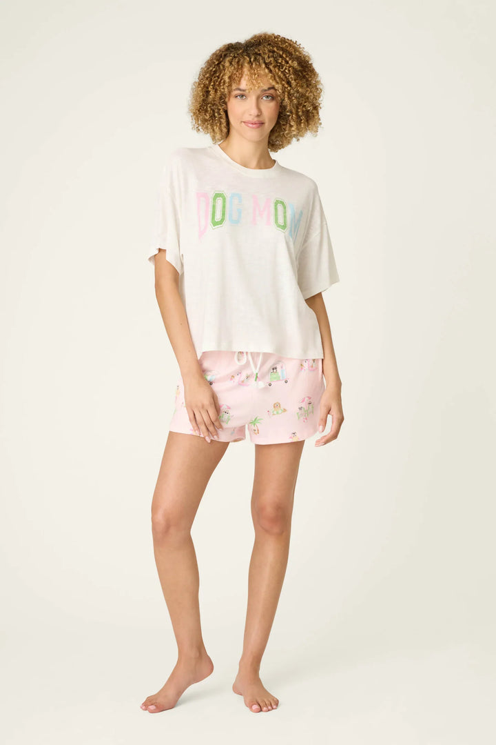 A woman with curly hair wears the Zertz PUPCATION PALMS DOG MOM TEE, a relaxed cotton-modal slub jersey with colorful lettering, paired with pink printed shorts and standing barefoot against a plain light background.