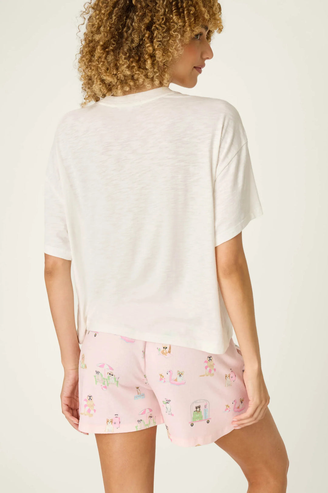 A woman with curly hair stands facing away from the camera, wearing the Zertz PUPCATION PALMS DOG MOM TEE—a relaxed crewneck made from cotton-modal slub jersey—and light pink, playfully patterned shorts.