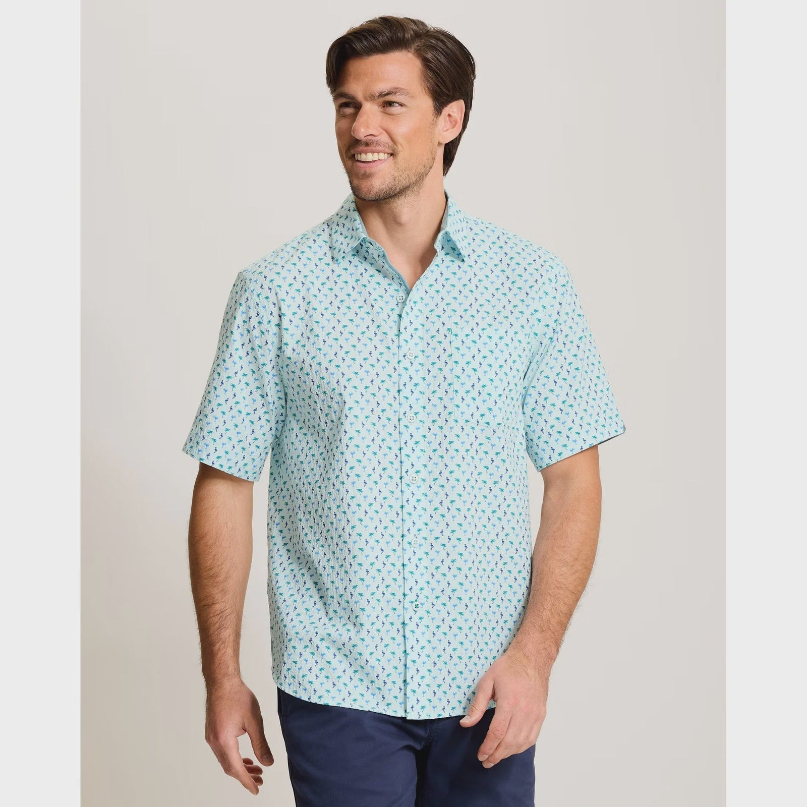 A man wears the Zertz NOVA WAVE FLOCK IT TO YOU light blue short-sleeve cotton shirt featuring a dark blue and red island-inspired print, paired with navy pants, standing and smiling against a plain light background.