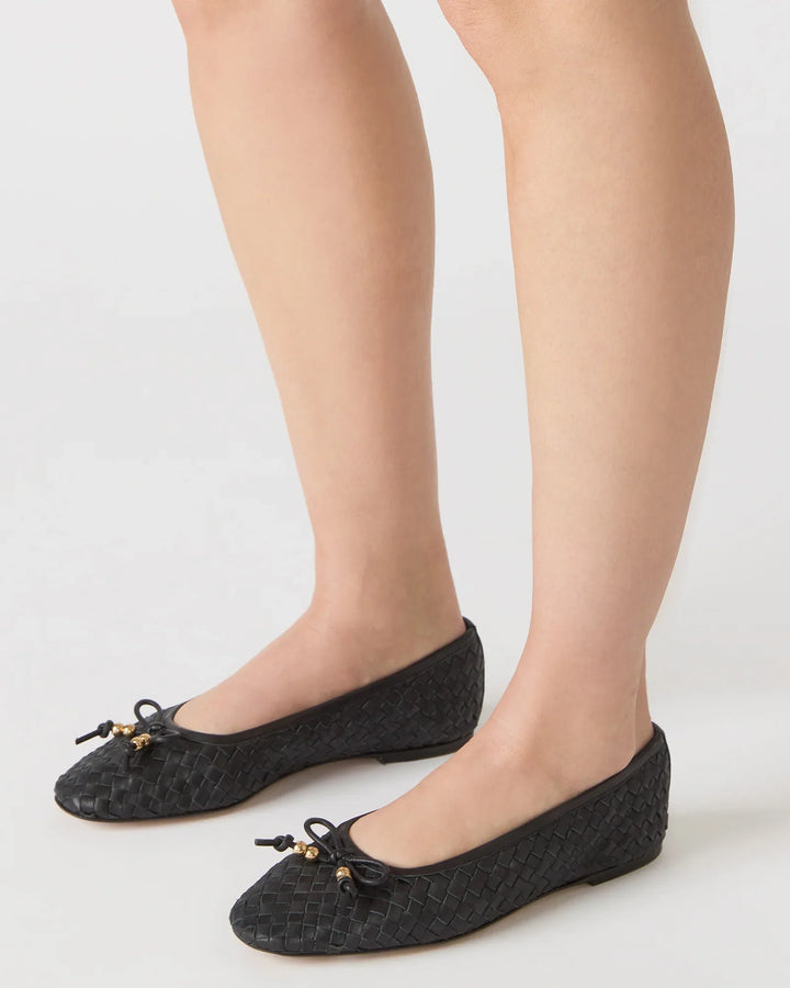 A person wears Zertz DIVY LEATHER FLATs—black woven ballet flats featuring small bows and gold charm accents. Only the lower legs and feet are shown, emphasizing the chic, embellished style against a white background.