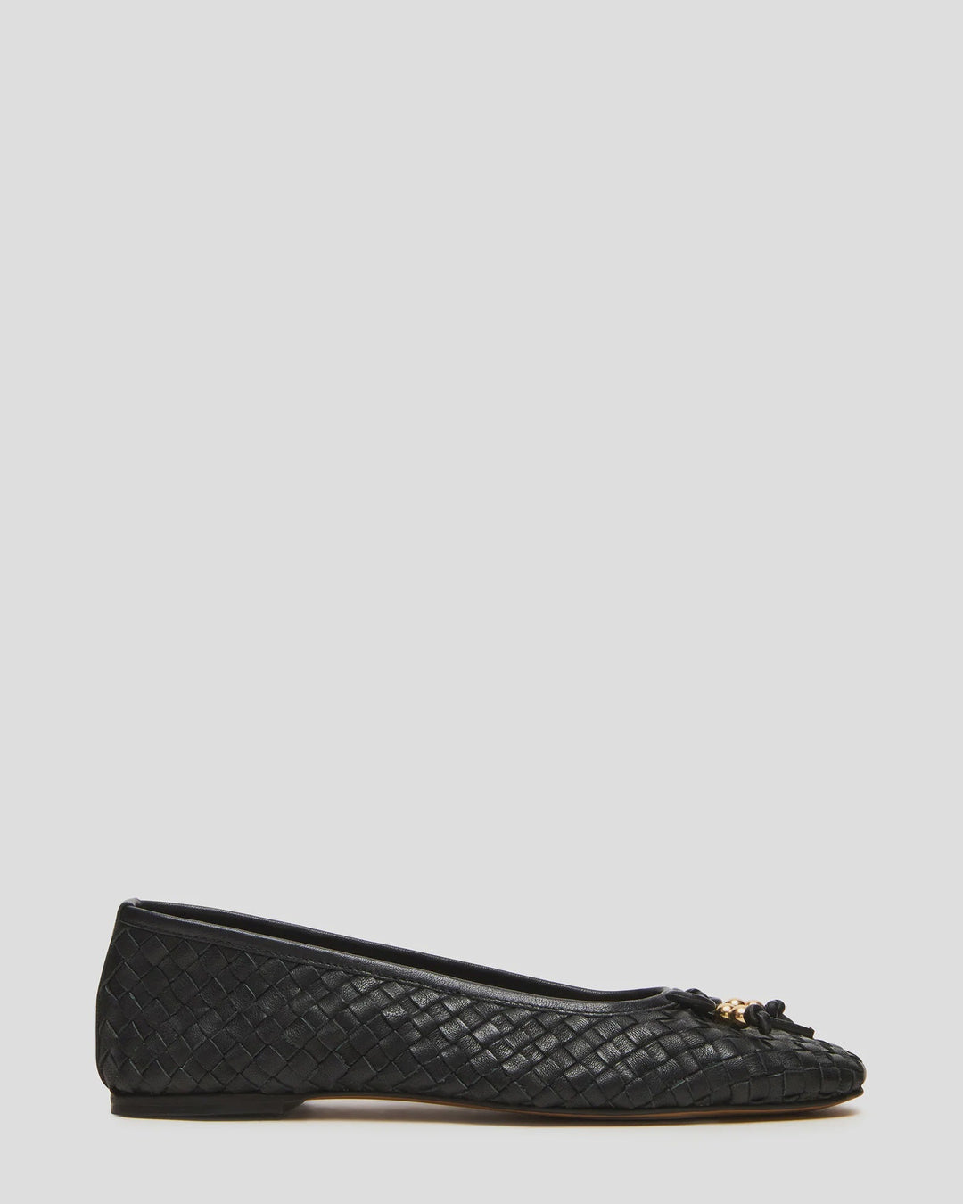 The Zertz DIVY LEATHER FLAT is a black leather shoe with a woven design, small bow, and gold-tone charm detail on the toe, shown in side profile against a light gray background. Ideal for those who love elegant, bow-embellished flats.