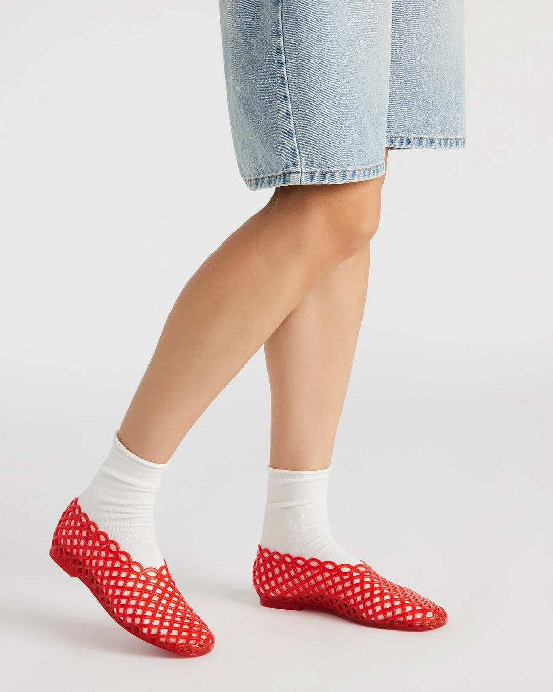 A person wearing knee-length light blue denim shorts, white socks, and Zertz GROVE JELLY SLIP ON red mesh ballet flats with a jelly cage design stands against a white background—ideal for everyday comfort.
