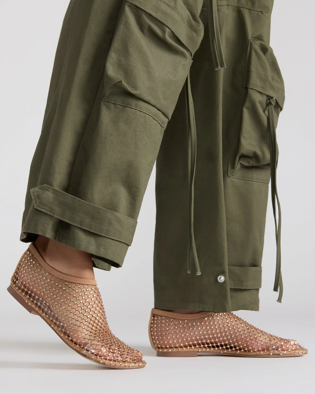 Wearing olive green cargo pants and Zertz’s MINO RHINESTONE FLAT, tan mesh flats adorned with rhinestones, a person stands against a plain background, highlighting an elegant style.