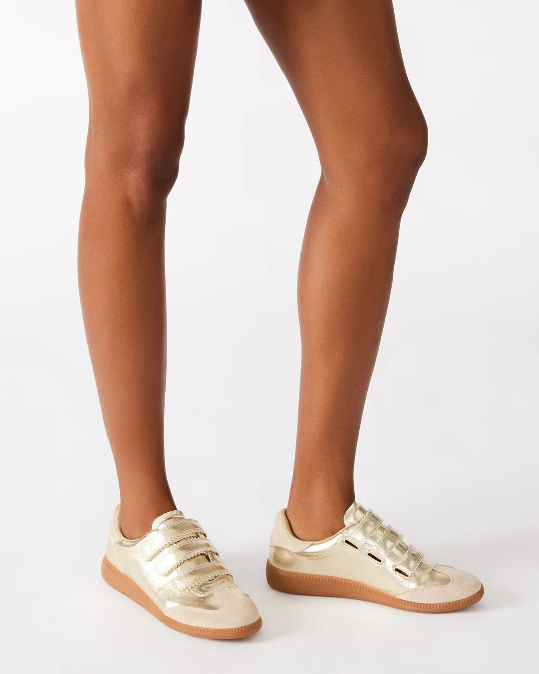 Wearing Zertz MOMENTUM SNEAKERS—shiny gold leather and suede low-tops with beige soles and Velcro straps—a person stands on a white background, legs visible from mid-thigh down.
