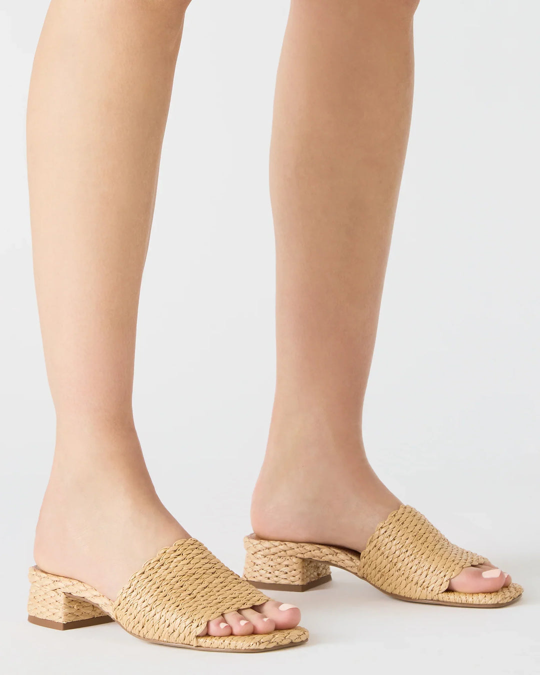 A person wears the Zertz MONTGOMERY SLIDE SANDAL, featuring a beige woven raffia upper, square toe, and open back, standing on a white surface.