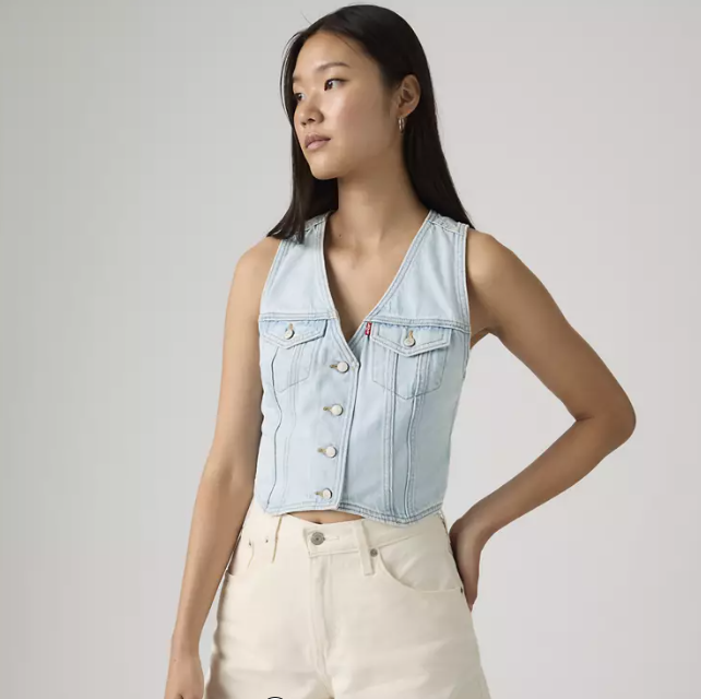 A woman with long straight hair stands against a plain background, wearing the Zertz Racer Vest - Never Going To Change in light blue and cream high-waisted pants. She looks off to the side with one hand on her hip.