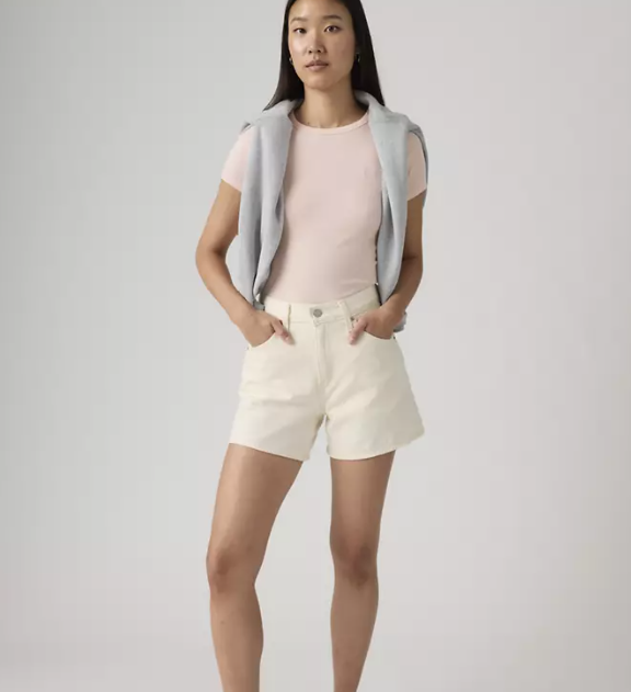 A woman stands confidently with her hands in the pockets of her Zertz CINCH MID-THIGH SHORT - BLANK IMPACT, styled with a light pink t-shirt and a light grey sweater over her shoulders against a plain background.