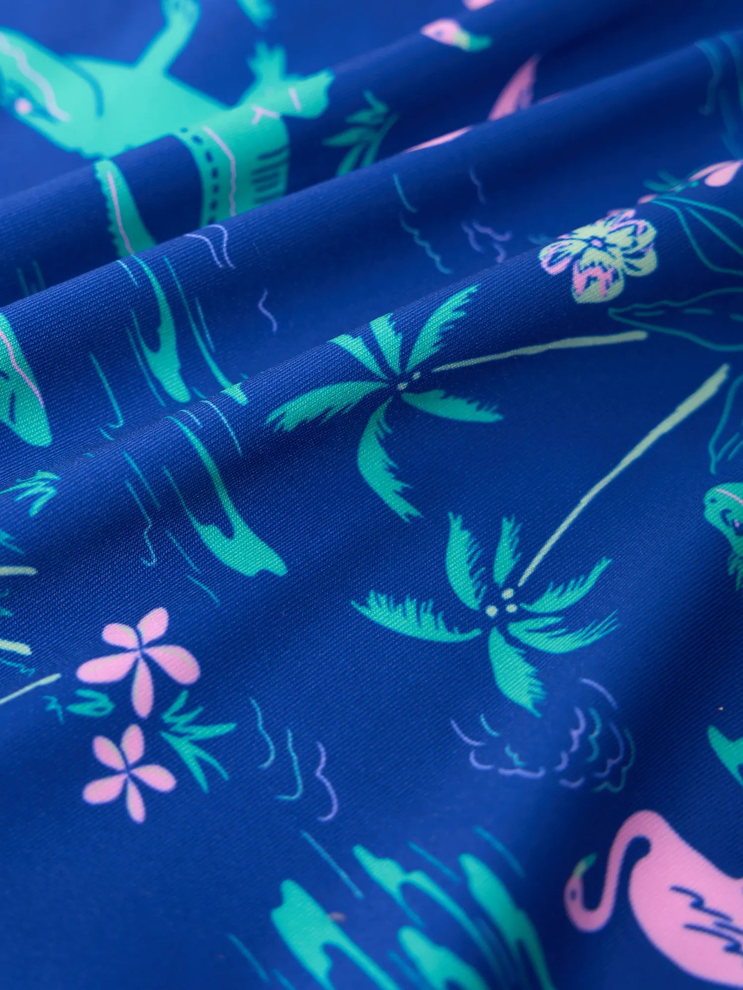 Close-up of the Zertz THE LIL SWAMP&CIRCUMSTANCE BOYS PERFORMANCE POLO, showcasing blue, stretchy UPF 50+ fabric with tropical green palms, pink flowers, flamingos, and alligators in a playful vibrant print.