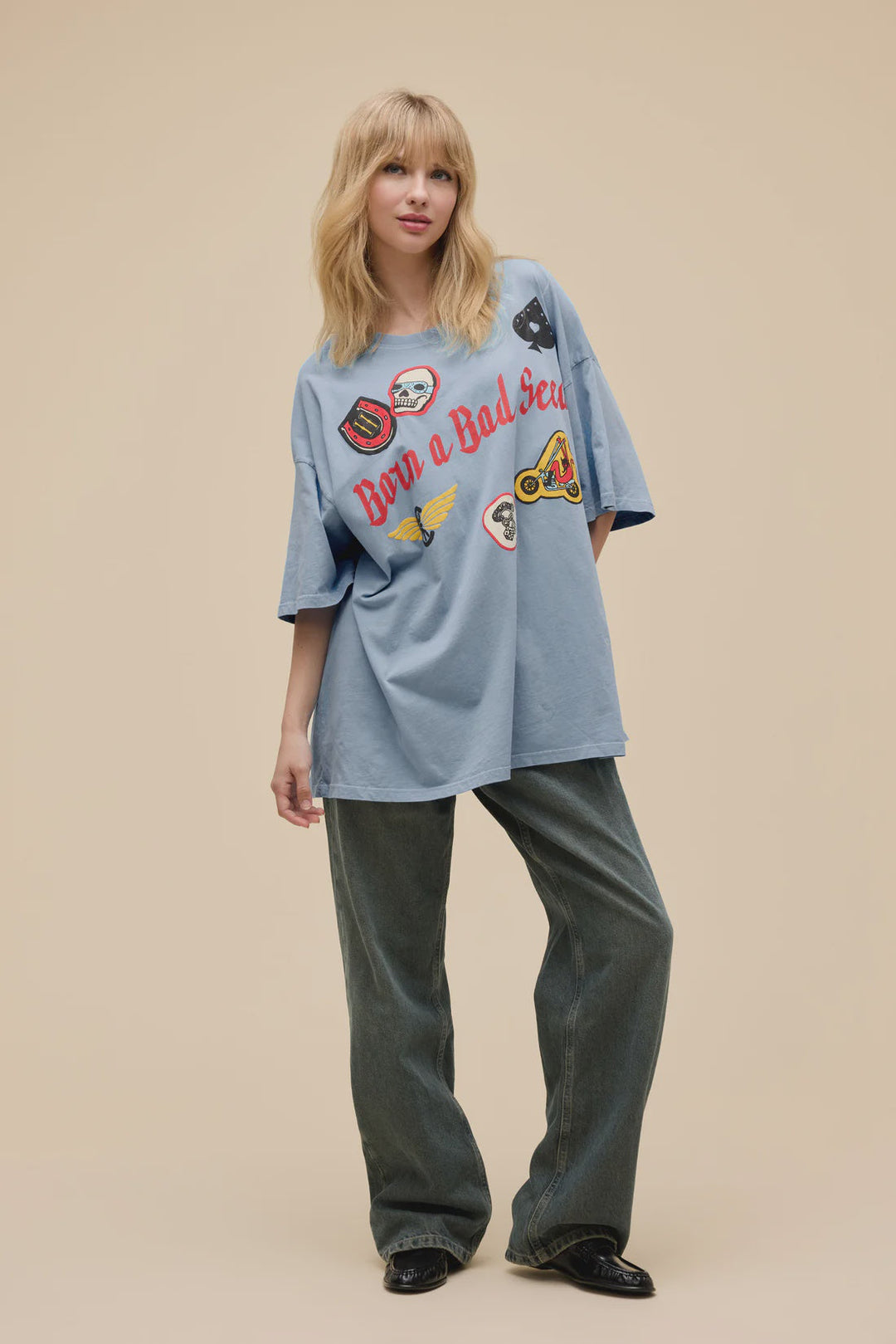 A blonde woman wears the Zertz BORN ON A BAD SEED OS TEE with puff ink patches, styled with wide-leg jeans and black shoes, standing before a plain beige background.