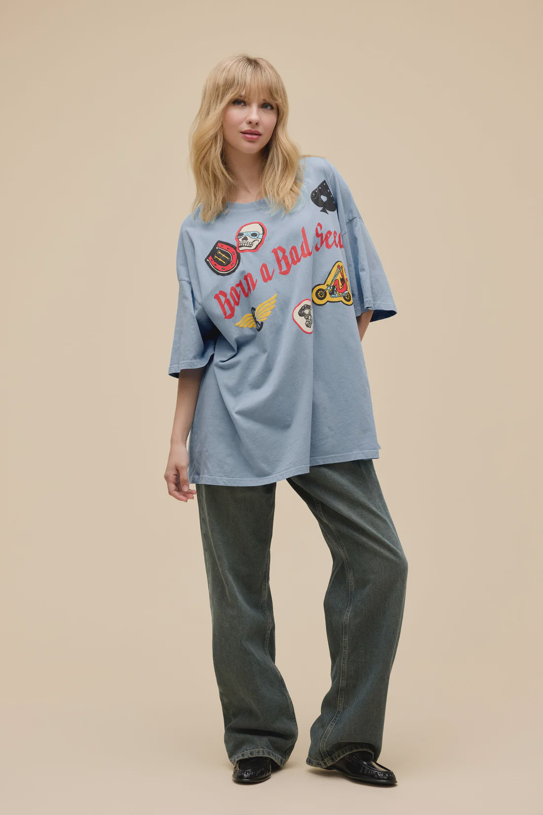 A blonde woman wears the Zertz BORN ON A BAD SEED OS TEE with puff ink patches, styled with wide-leg jeans and black shoes, standing before a plain beige background.