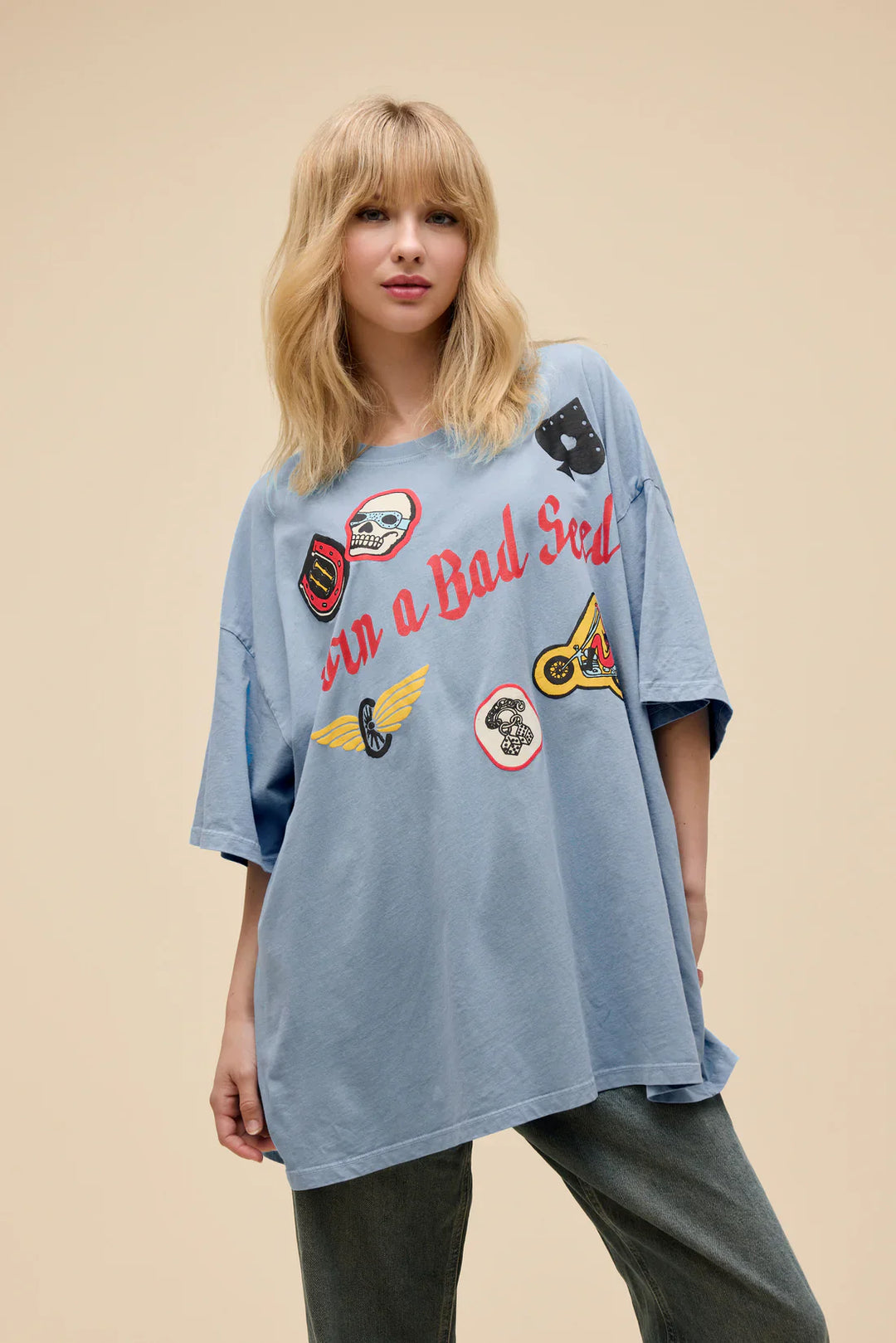 A woman with blonde hair models the Zertz BORN ON A BAD SEED OS TEE in light blue, featuring vibrant puff ink patches and bold red lettering, against a plain beige background.