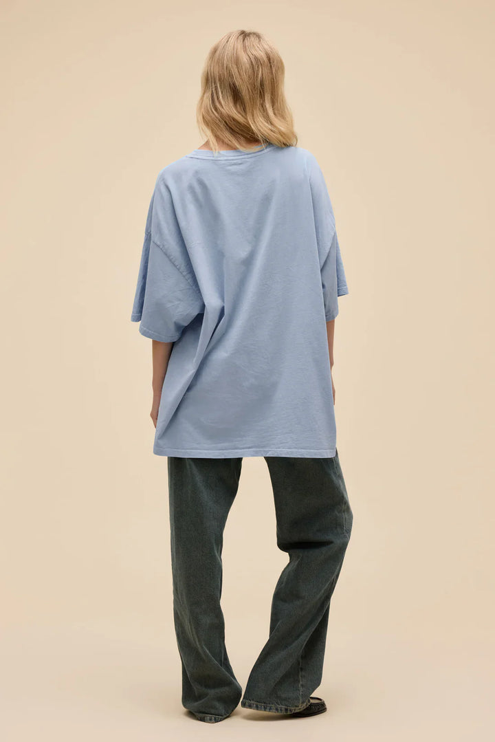 A person with long blonde hair stands facing away from the camera, wearing a loose Zertz BORN ON A BAD SEED OS TEE and dark wide-leg jeans with puff ink patches, set against a beige background.
