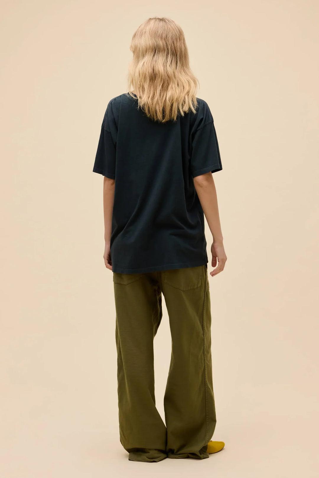 A person with wavy blonde hair stands facing away, wearing a loose dark CHRIST STAPLETON EAGLE MERCH TEE by Zertz, wide olive green pants, and yellow shoes against a plain beige background.