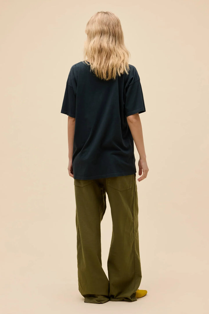 A person with wavy blonde hair stands facing away, wearing a loose dark CHRIST STAPLETON EAGLE MERCH TEE by Zertz, wide olive green pants, and yellow shoes against a plain beige background.