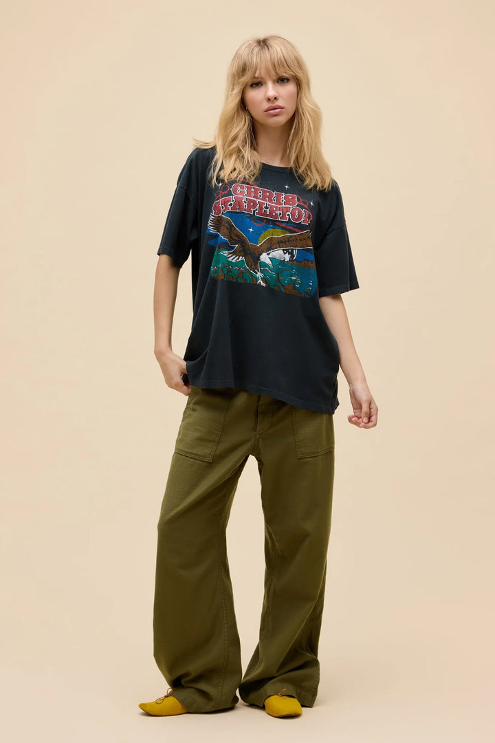 A blonde woman wears the Zertz CHRIST STAPLETON EAGLE MERCH TEE in black, styled with olive green wide-leg pants and yellow shoes, standing against a plain beige backdrop—ideal for any country collection.