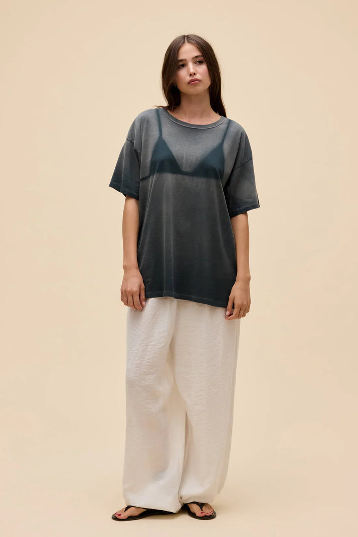 A woman with long brown hair poses in front of a beige background wearing the Zertz WET BIKINI MERCH TEE—a dark, semi-sheer t-shirt with a sun-faded print over a black bra—styled with wide white pants and black sandals.