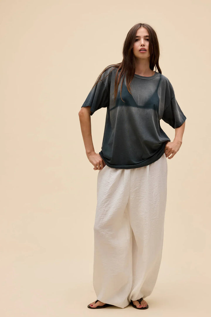 Against a beige background, a woman models the Zertz WET BIKINI MERCH TEE over a black bra, paired with loose white wide-leg pants and black sandals. Her long brown hair is down as she poses casually with one hand on her hip.