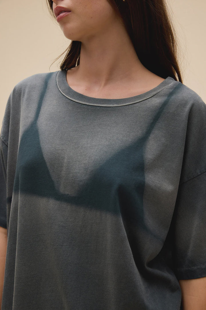 A woman with long dark hair models the Zertz WET BIKINI MERCH TEE. The sun-faded print and her bra outline are faintly visible through the loose fabric. Image is cropped from nose to mid-torso against a neutral background.