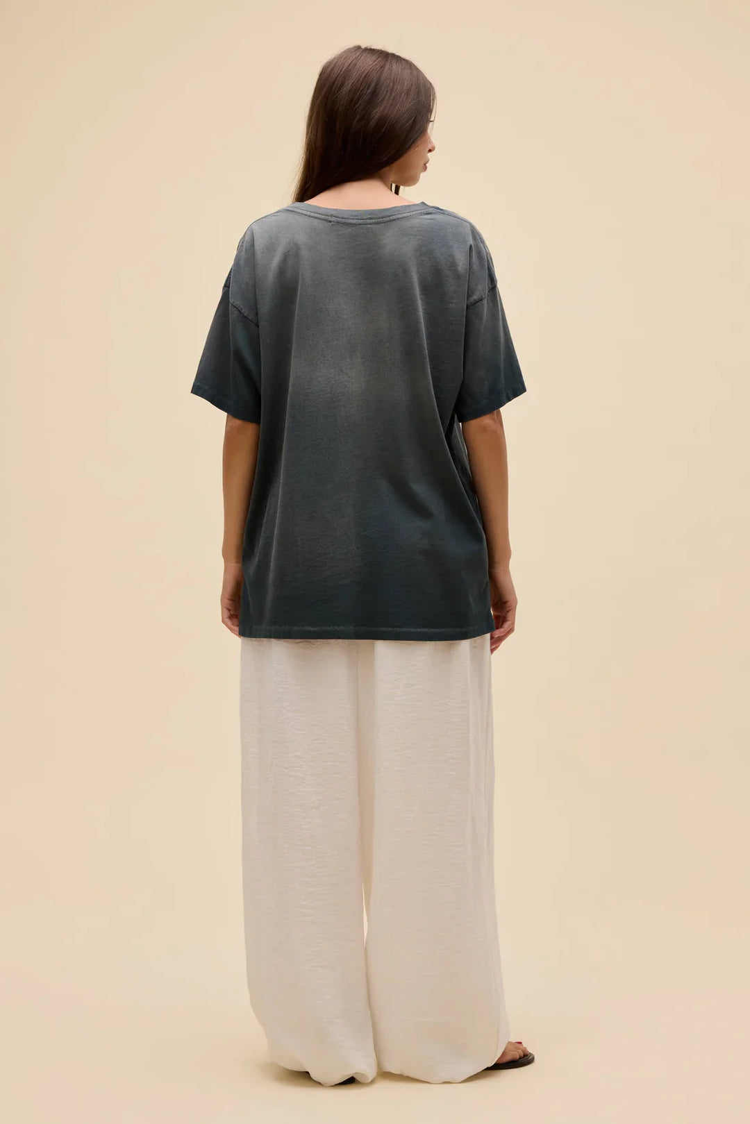 A woman with long brown hair stands against a beige background, wearing the Zertz WET BIKINI MERCH TEE featuring a sun-faded print and loose fit, paired with wide-leg white pants.