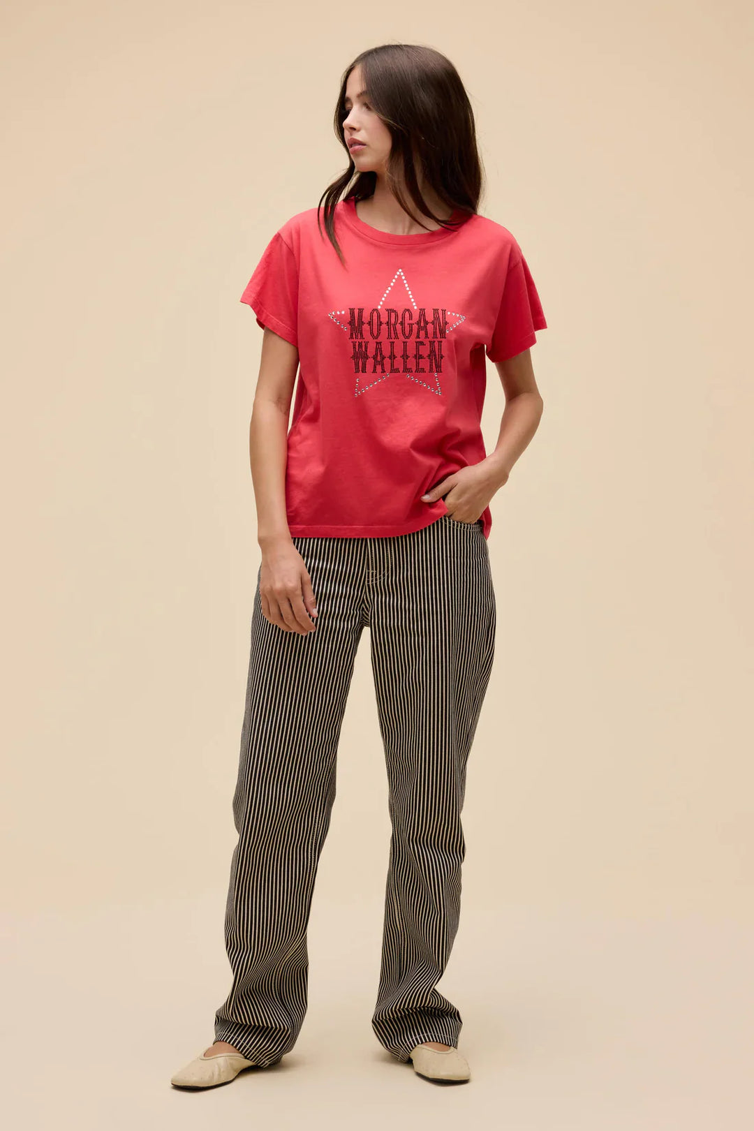 A woman with long brown hair stands against a beige background wearing the Zertz MORGAN WALLEN RHINESTONE STAR SOLO TEE and black-and-white striped pants, looking to the side with one hand in her pocket.
