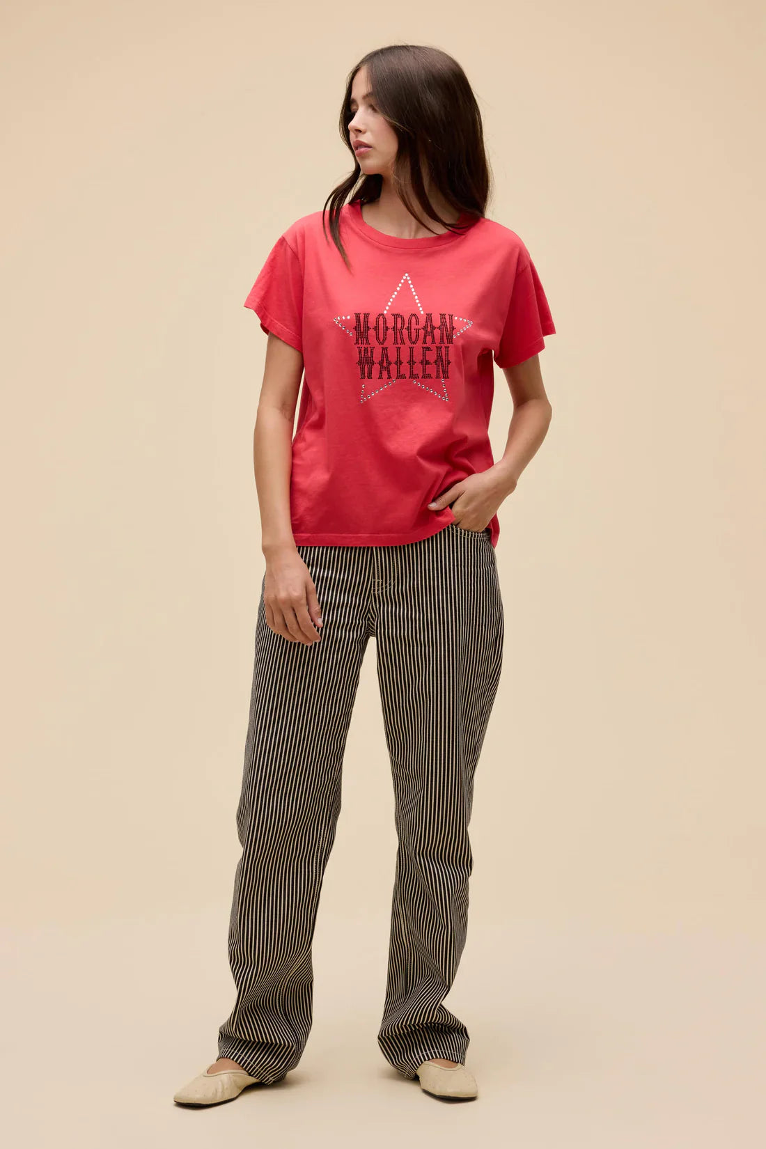 A woman with long brown hair stands against a beige background wearing the Zertz MORGAN WALLEN RHINESTONE STAR SOLO TEE and black-and-white striped pants, looking to the side with one hand in her pocket.