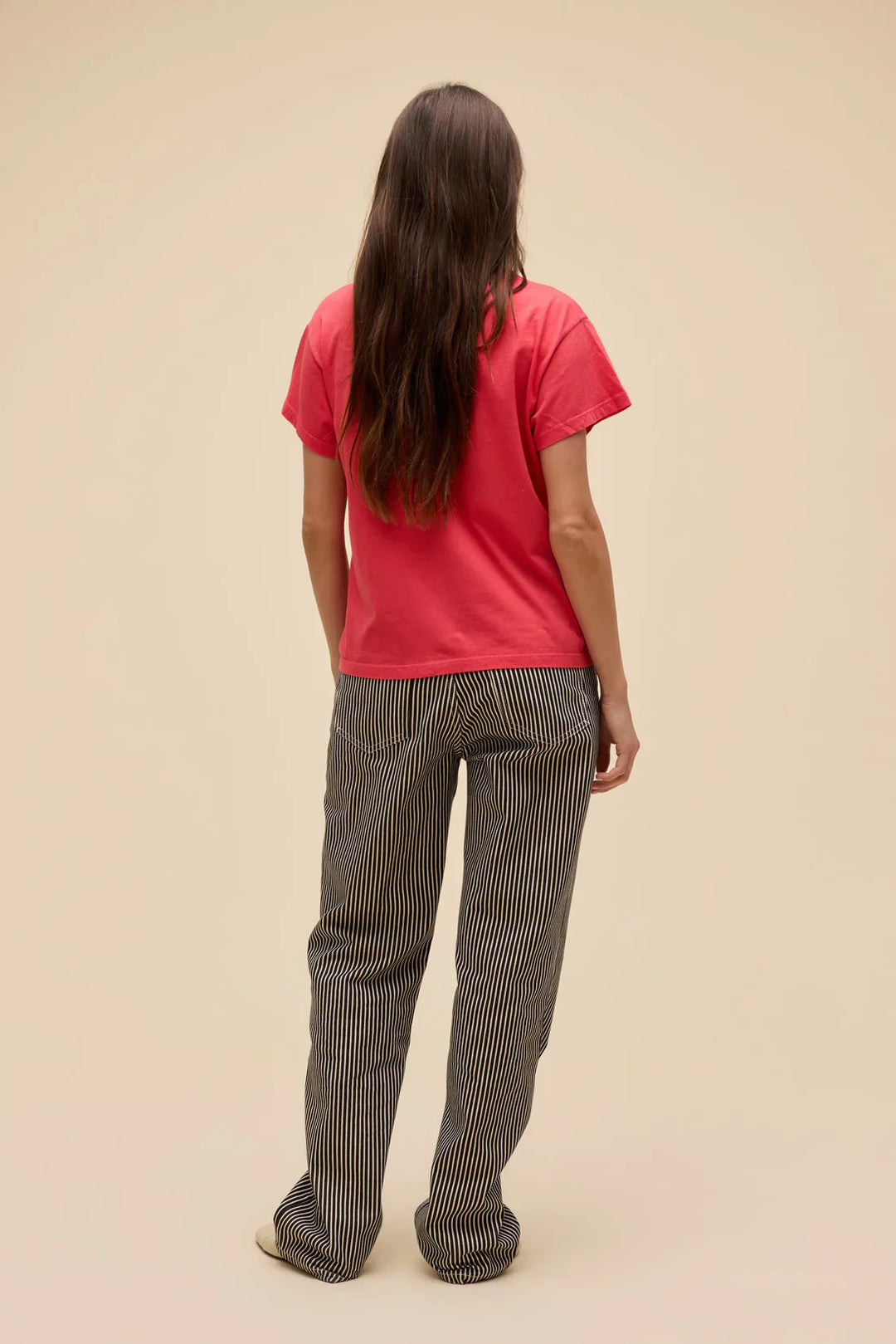 A person with long brown hair faces away against a plain beige background, wearing the Zertz MORGAN WALLEN RHINESTONE STAR SOLO TEE in red with western lettering and black-and-white striped pants.