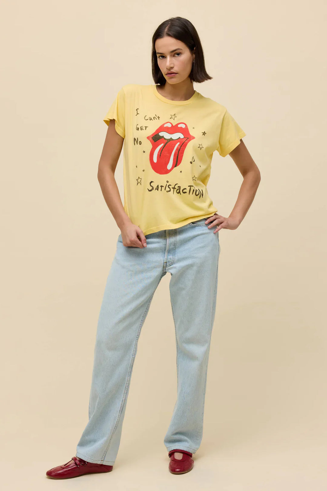 A woman with straight brown hair wears the Zertz ROLLING STONES SATISFACTION DOODLES SOLO TEE in yellow, light blue jeans, and red shoes, standing with one hand in her pocket against a beige background.
