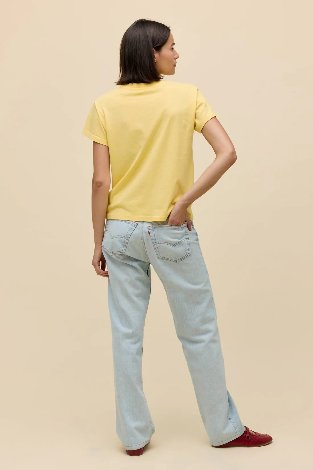 A person with short brown hair, wearing the Zertz ROLLING STONES SATISFACTION DOODLES SOLO TEE, light blue jeans, and red shoes stands facing away with one hand in their back pocket against a plain beige background, looking satisfied.