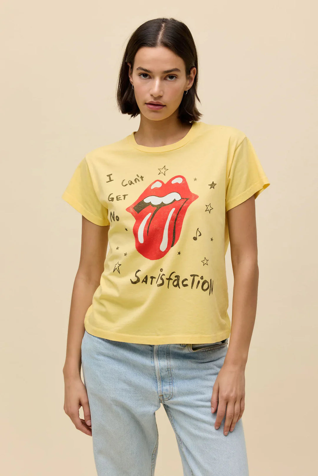 A woman wears the Zertz ROLLING STONES SATISFACTION DOODLES SOLO TEE—a yellow shirt with the band’s classic tongue logo—paired with light blue jeans, standing in front of a plain beige background.