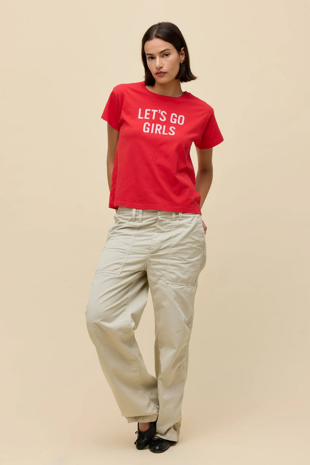 A woman stands against a beige background wearing the Zertz SHANIA TWAIN LETS GO GIRLS SOLO TEE in red, paired with light beige cargo pants and black shoes, her hands in her pockets.