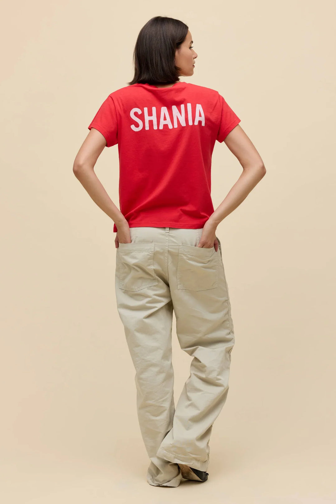 A person stands facing away, wearing the Zertz SHANIA TWAIN LETS GO GIRLS SOLO TEE with "SHANIA" in white on the back and light pants, hands in pockets, against a beige background. Let&