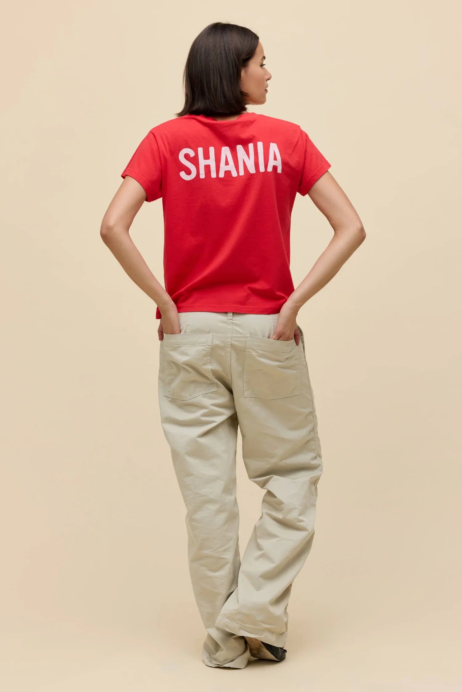 A person stands facing away, wearing the Zertz SHANIA TWAIN LETS GO GIRLS SOLO TEE with "SHANIA" in white on the back and light pants, hands in pockets, against a beige background. Let's Go Girls!.