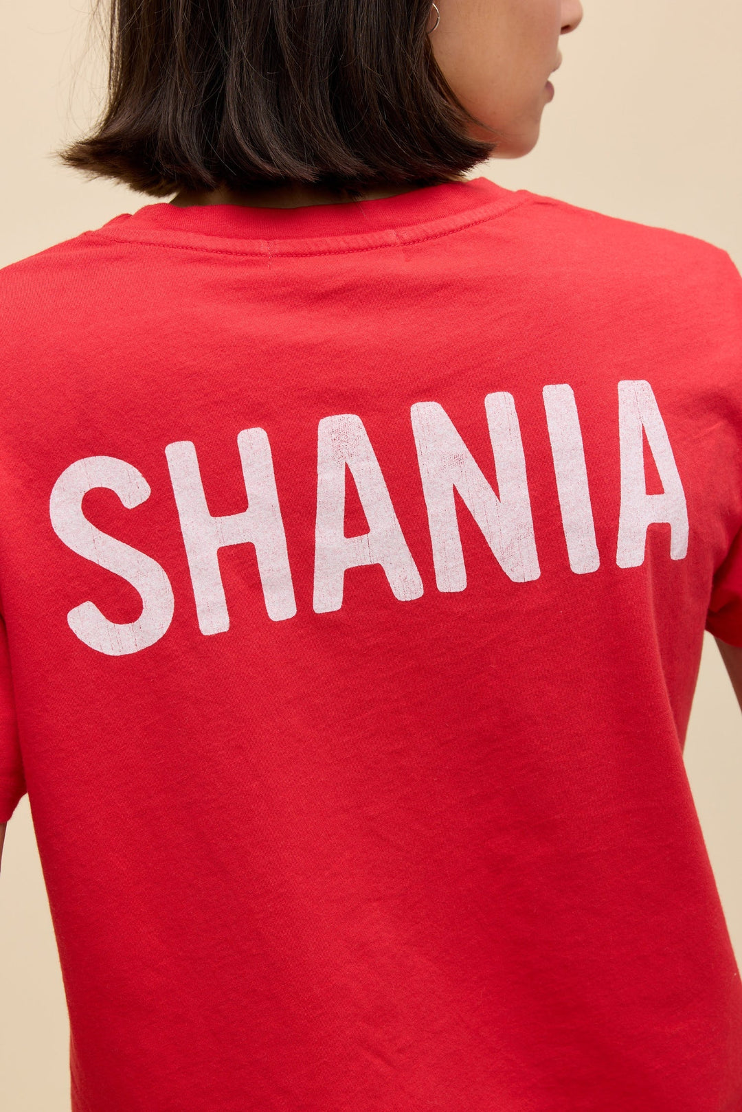 A person wears the Zertz SHANIA TWAIN LETS GO GIRLS SOLO TEE in red, featuring bold white SHANIA lettering on the back. The photo captures their upper back and head from behind, radiating true "Let&