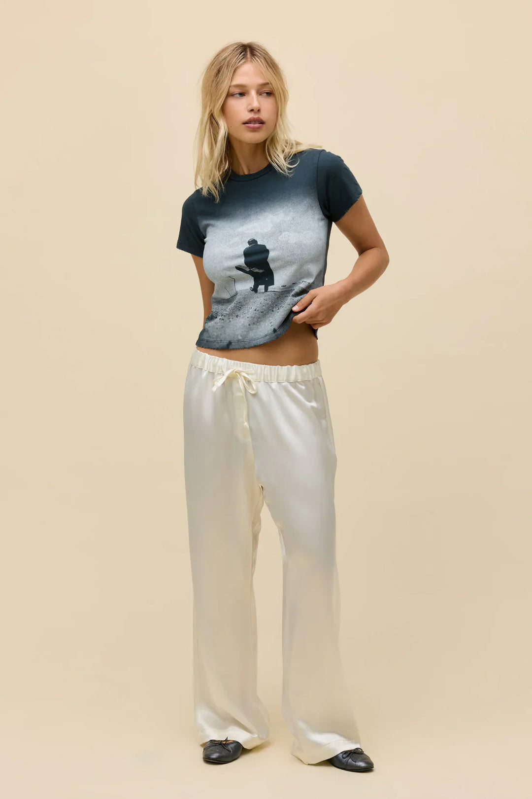 A woman stands against a beige background wearing the Zertz JOHNNY CASH SILHOUETTE VNTG TEE, cream satin drawstring pants, and black shoes. She looks to the side, holding the hem of her shirt with one hand.